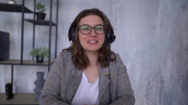 Young attractive woman is recording a video tutorial. Girl in glasses and in headphones with a microphone speaks smiling and looking at the camera