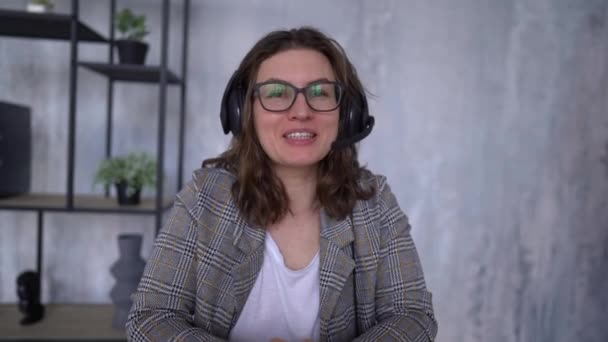 Young attractive woman is recording a video tutorial. Girl in glasses and in headphones with a microphone speaks smiling and looking at the camera — Video Stock