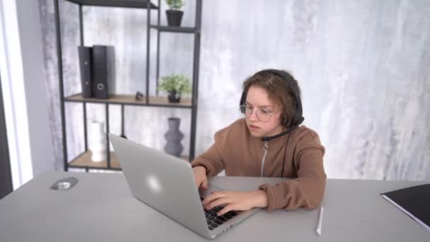 Portret uroczej nastolatki, która ma video-lekcję online. Dziewczyna siedzi przy laptopie ze słuchawkami, słucha i pisze. Szczęśliwa młoda dziewczyna uczyć się języka angielskiego słuchać wykładu — Wideo stockowe