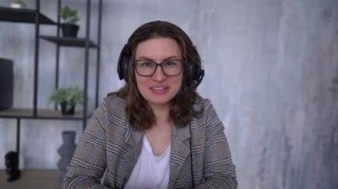 Young beautiful caucasian woman with wireless headphones speaks in a video call greeting and looking at the camera in a white living room in elegant style. Remote communication concept