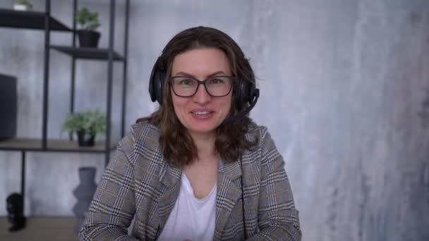 Young beautiful caucasian woman with wireless headphones speaks in a video call greeting and looking at the camera in a white living room in elegant style. Remote communication concept — Stockvideo