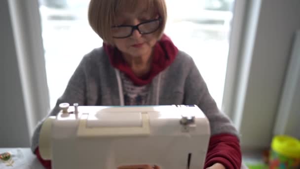 Rentnerin mit Brille näht Heimtextilien an einer Nähmaschine. Rentnerin und ihr Hobby, Großmutter arbeitet zu Hause — Stockvideo