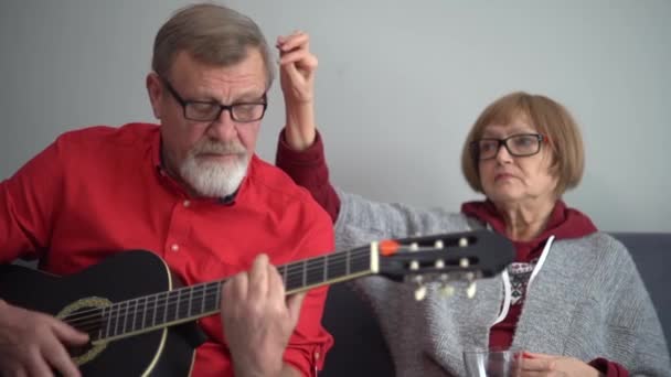 Amantes pareja de ancianos, marido y mujer, los pensionistas pasan tiempo juntos sentados en el sofá. Un hombre con camisa roja toca la guitarra acústica y canta para su esposa — Vídeo de stock