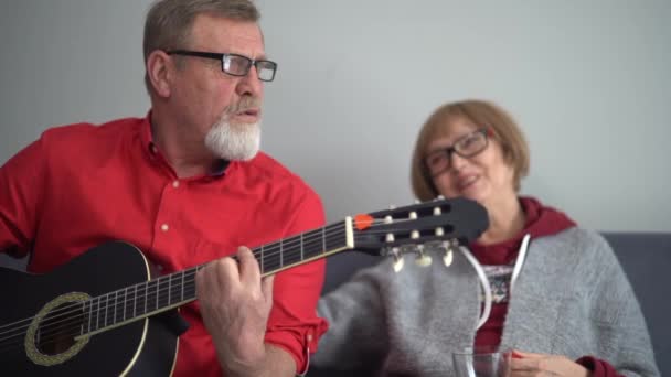 Man in a red shirt plays the acoustic guitar and sings for his wife. Loving elderly couple, husband and wife, pensioners spend time together sitting on the couch — 비디오