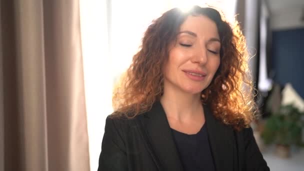 Curly business woman in a black business suit looking at the camera while smiling while standing in front of the window in the office — Stockvideo