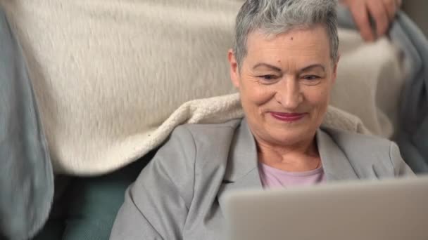 一个有着灰色短发的成熟女人的近照是坐在沙发上与笔记本电脑一起工作。她有爱心的丈夫用毯子盖住他的妻子 — 图库视频影像