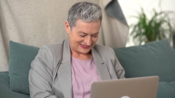 Mulher madura com um corte de cabelo curto cinza está trabalhando sentado no sofá com um laptop. Seu marido carinhoso cobre sua esposa com um cobertor — Vídeo de Stock