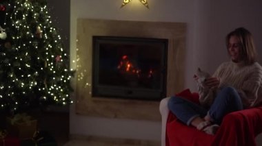 Güzel bir kadın şöminenin yanındaki koltukta otururken sıcak çoraplar giyer ve yılbaşı ağacı süslenir. Mutlu Noeller.