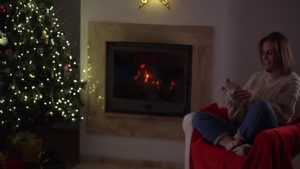 Mulher bonita coloca meias quentes enquanto sentado em uma poltrona junto à lareira e árvore de Natal decorada Ano Novo. Feliz Natal! — Vídeo de Stock
