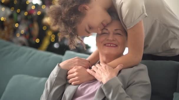 Šťastná babička s krátkým sestřihem a šedivými vlasy sedí na gauči. Kudrnatý chlapec vnuk teenager objetí ji na pozadí vánočního stromečku — Stock video