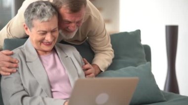 Mutlu yaşlı çift gülüyor, konuşuyor, uzaktan video görüşmesi yapıyor, dizüstü bilgisayara, webcam 'e bakıyor, kanepede oturuyor, neşeli olgun yaşlı aile evde internetten sohbet ediyor.