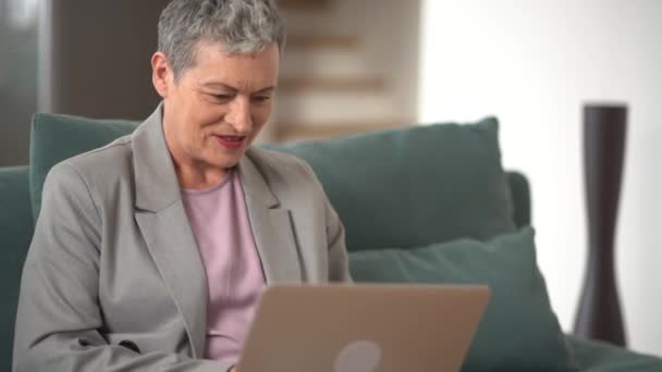 视频呼叫概念，可爱的老年妇女，有着短短的灰发，面带微笑，与家人聊天，用笔记本电脑进行视频连接，远程见面，打招呼，高兴地看到，坐在沙发上 — 图库视频影像