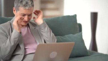 Gri takım elbiseli ve kısa gri saçlı olgun bir kadın dizüstü bilgisayarla video görüşmesi yapıyor ve evdeki kanepede oturuyor. Mutlu emekli kadın konsepti