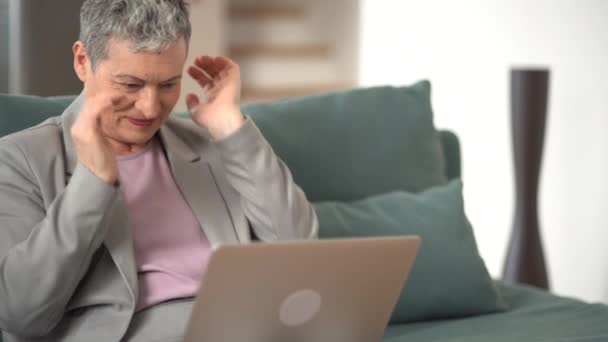 Starší žena v šedém obleku a s krátkými šedými vlasy má videohovor pomocí notebooku a sedí doma na gauči. Šťastná žena v důchodu koncept — Stock video