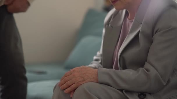 Θλιβερή ηλικιωμένη γυναίκα κάθεται στον καναπέ στο σπίτι και ο σύζυγός της έρχεται και παίρνει τα χέρια της. Κοντινό πλάνο του χεριού ενός ώριμου ηλικιωμένου ζευγαριού, έννοια στήριξης και τρυφερότητας — Αρχείο Βίντεο