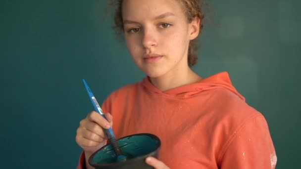 Retrato de perto de uma linda garota encaracolada com um pincel e uma tigela de tinta azul. Criança mesmo faz reparos em seu próprio quarto, pinta parede com tinta teal. Conceito de renovação ou redecoração de casa — Vídeo de Stock