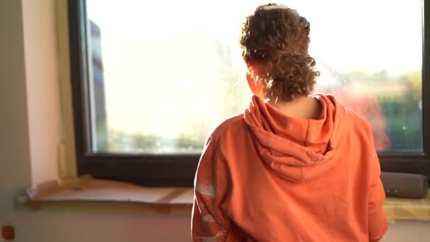 Retrato alegre de uma adolescente ruiva encaracolada em um capuz manchado de tinta laranja na frente de uma janela em uma casa a ser renovada — Vídeo de Stock