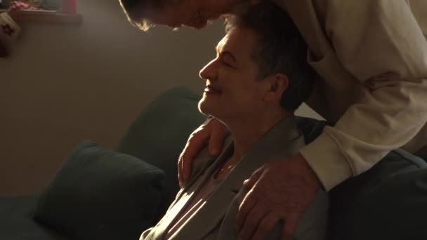 Heerlijk ouder liefhebbend koppel. De man knuffelt zijn vrouw zittend op de bank van achteren en kust haar op het voorhoofd, een gelukkig gezin en trouw — Stockvideo