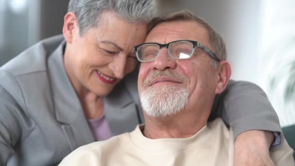 Hermosa pareja de cabello gris abrazándose mientras está sentada en el sofá. Una mujer con un corte de pelo corto con estilo y un hombre con barba y gafas. Feliz concepto de vejez — Vídeos de Stock