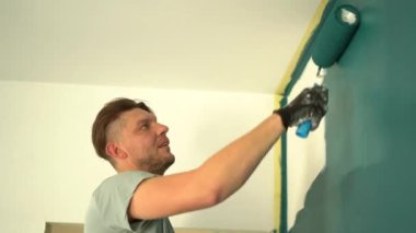 Yan görüş, alçak açı. Genç bir adam yeni evinden bir boya silindiriyle duvar boyuyor. Emlak ve yenileme konsepti