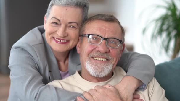 Hombre y una mujer abrazándose y mirando a la cámara mientras están sentados en el sofá. Indore retrato de una pareja de ancianos casados de pelo gris — Vídeos de Stock