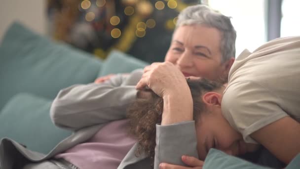 Großmutter und Enkel feiern Weihnachten. Schöne Teenager-Junge mit lockigem Haar umarmt eine stilvolle grauhaarige Frau sitzt auf einem Sofa vor dem Hintergrund der Weihnachtsbaumbeleuchtung — Stockvideo