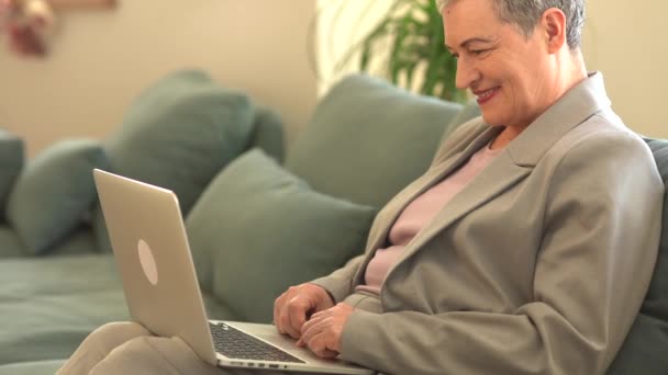 Krásná šedovlasá babička s krátkým účesem v šedém obleku sedí doma na pohovce v obývacím pokoji. Koncept vzdálené zaměstnanosti, aktivního odchodu do důchodu a zralého věku — Stock video