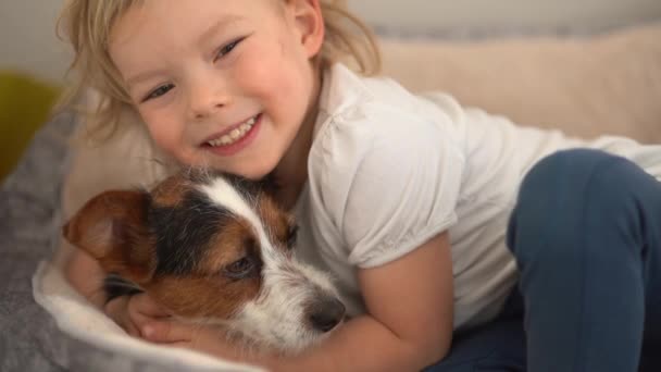 Śmiejący się chłopiec leży na psim łóżku obok swojego małego zwierzaka, kundla. Cute trzyletnie dziecko i jego ukochanego psa — Wideo stockowe
