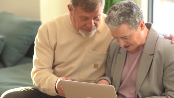 Retrato de una pareja de ancianos de pelo gris sentados en un sofá con un portátil en el regazo y una tarjeta bancaria en las manos. Jubilación activa y concepto de compras en línea — Vídeos de Stock