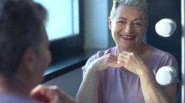 Gri saçlı, gülümseyen yaşlı bir kadının yakın plan fotoğrafı, cilt bakımı ve makyaj. Mutlu, çekici, yaşlı kadın aynada kendine bakıyor.