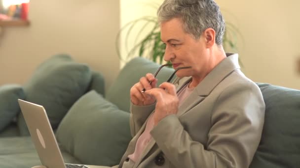 Empleo remoto, una freelancer con un corte de pelo corto gris mira a la cámara trabajando con un portátil mientras está sentada en el sofá en casa — Vídeos de Stock