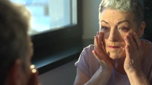 Beautiful senior woman checking her face skin and looking for blemishes. Wrinkled lady with grey hair checking wrinkles around eyes, aging process. Happy senior woman looking in the mirror. Close-up — Stock Video