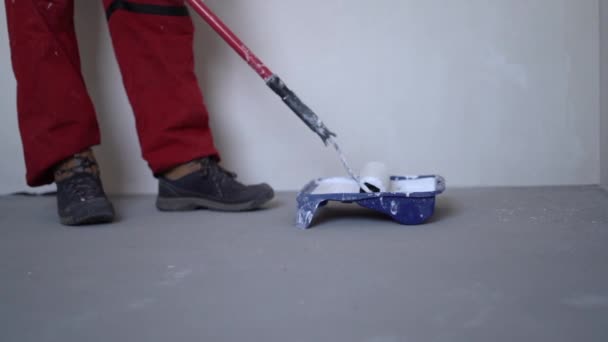 A gray-haired mature man in a red overalls dunks a paint roller into a pallet of white paint. Repair in a new house concept — Stock Video