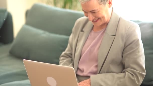 Sonriente dama de pelo gris en un traje de negocios está trabajando con un ordenador portátil en casa solo mientras está sentado en el sofá. Concepto de empleo remoto, freelancer femenina, jubilación feliz — Vídeos de Stock