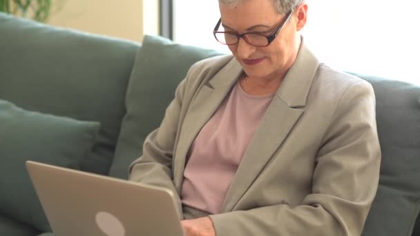 Actief gelukkig senior vrouw draagt bril en kort kapsel zit op de bank en werkt op afstand met behulp van laptop — Stockvideo