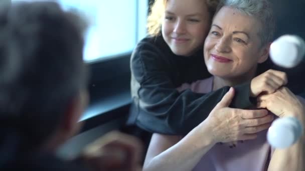 Nastoletnia dziewczyna przytula swoją piękną, siwą babcię siedzącą przy lustrze. Dwa pokolenia jednej rodziny, zmiany związane z wiekiem i koncepcja starzenia się — Wideo stockowe