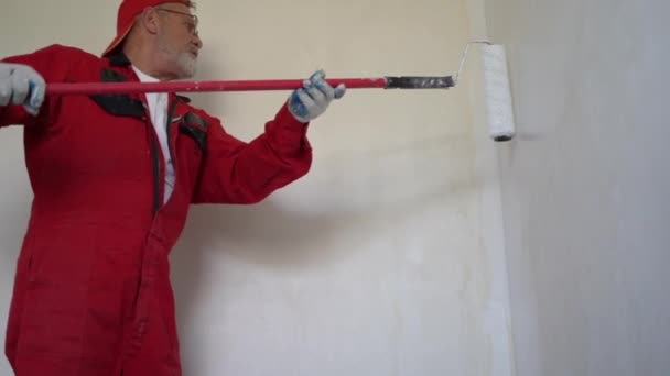 Een bejaarde gepensioneerde in een rode overall repareert zijn huis. Een man schildert een muur in slechte kleur met een roller op een lange steel — Stockvideo