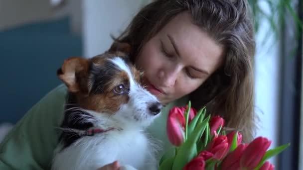 Усміхнена біла жінка з букетом тюльпанів цілує свого собаку, яка сидить на підлозі вдома. спосіб життя, домашня тварина, товариство та концепція дружби тварин — стокове відео