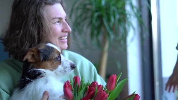 Feliz madre en el día de las madres sostiene un perro y un ramo de tulipanes en sus manos mientras su pequeño hijo la abraza. Cumpleaños niña o día internacional de las mujeres — Vídeo de stock