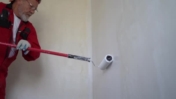 Een man schildert een muur in slechte kleur met een roller op een lange steel. Een bejaarde gepensioneerde in een rode overall repareert zijn huis — Stockvideo