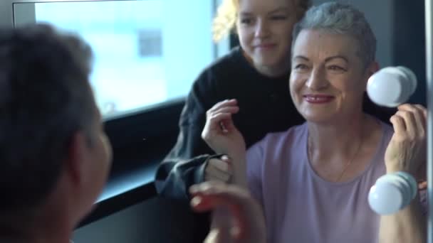 Зріла білошкіра жінка перевіряє зморшки очей перед дзеркалом. Портрет щасливої бабусі та юної онучки. Двоє поколінь однієї сім'ї — стокове відео