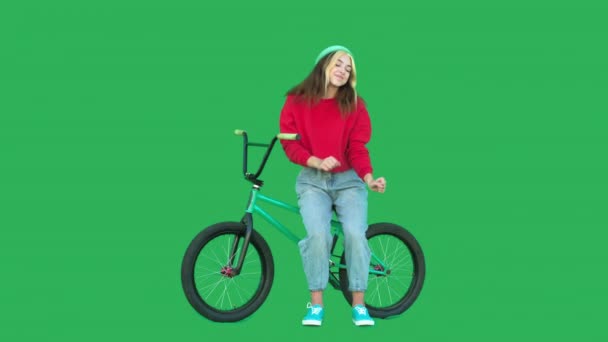 スウェットシャツとニット帽を身に着けているスポーティガールは 緑の画面の背景にBmxバイクで立っている 若い女性が自転車の近くでカメラを見て踊っている クロマキー 4K生ビデオ映像 — ストック動画