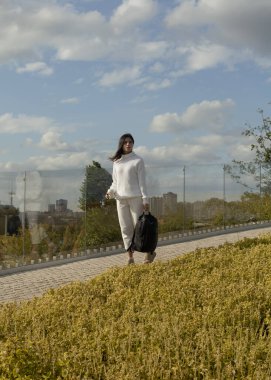 Beyaz sonbahar elbiseli bir kız şehir parkında sırt çantasıyla yürüyor.