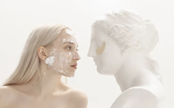 Yüzünde makyajlı bir kız hafif arka planda sıva heykeliyle poz veriyor.