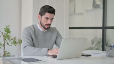 Ofiste dizüstü bilgisayar kullanırken kamerayı gösteren genç adam.
