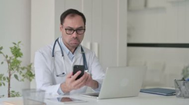 Ofiste dizüstü bilgisayar üzerinde çalışırken akıllı telefon kullanan orta yaşlı bir doktor. 