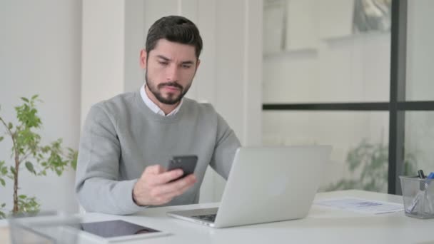 Wütender Geschäftsmann spricht auf Smartphone, während er Laptop im Büro benutzt — Stockvideo