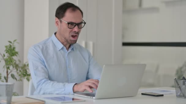 Чоловік середнього віку відчуває шок під час використання ноутбука в офісі — стокове відео