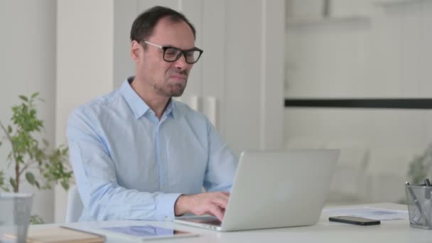 Людина середнього віку, яка відчуває біль у спині, користуючись ноутбуком у офісі — стокове відео