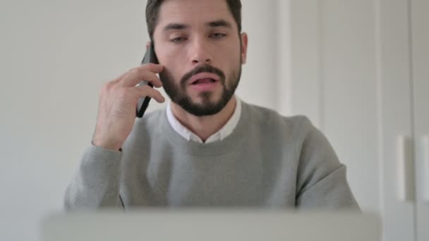 Porträt eines jungen Mannes, der mit dem Laptop auf dem Smartphone spricht — Stockvideo